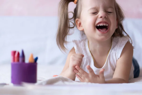 美しい女の子はベッドの中で鉛筆で描きます。2年生の少女がベッドの上に横たわる本のぬり絵を描く。ベッドの上に寝そべっている間、ポニーテールの笑みを浮かべて小さな赤ちゃん。きれいな女の子は鉛筆で描くと — ストック写真