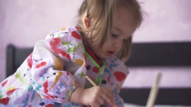 3 yaşındaki küçük bir kız bir kağıt parçasına resim yapıyor. Slav görünümlü güzel bir kız yatağın üzerinde oturuyor. — Stok video