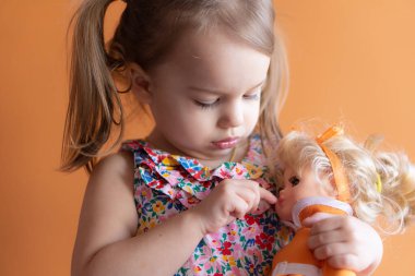 Turuncu arka planda üç yaşında güzel bir kızın portresi. Oyuncak bebekle oynayan çocuk