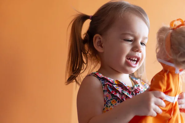 Portret van een mooi meisje van drie jaar op een oranje achtergrond. Kind spelen met pop — Stockfoto