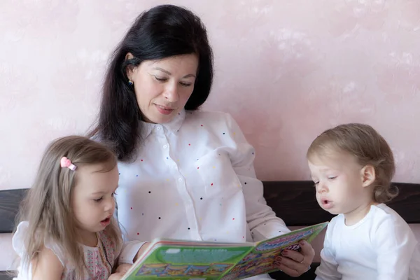 Die junge Großmutter liest ihren kleinen Enkeln ein Buch vor. Die junge schöne Großmutter verbringt Zeit mit ihren Zwillingsenkeln. Großmutter mit ihren Enkeln beim Lesen eines Buches im Bett — Stockfoto