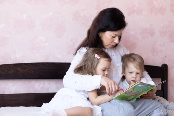 Die junge Großmutter liest ihren kleinen Enkeln ein Buch vor. Die junge schöne Großmutter verbringt Zeit mit ihren Zwillingsenkeln. Großmutter mit ihren Enkeln beim Lesen eines Buches im Bett — Stockfoto