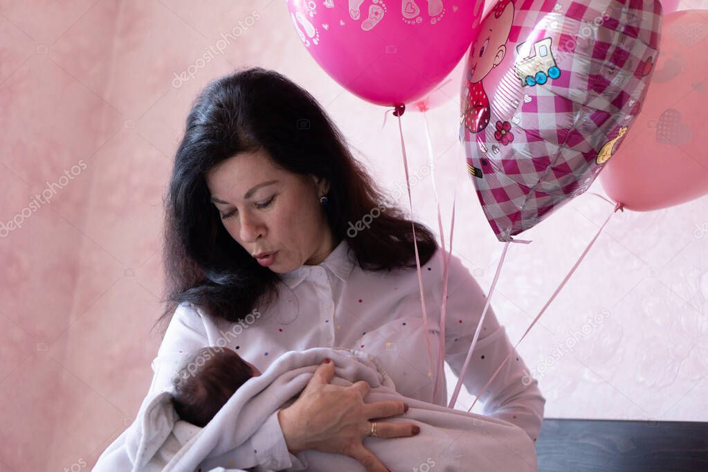 Young grandmother holding baby sitting on bed. Young beautiful grandmother spends time with newborn sitting in bed holding balloons in the hand