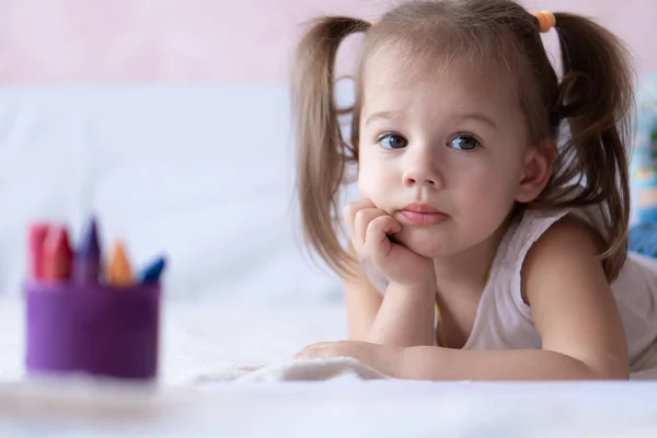 Kleines Mädchen von zwei Jahren malt Malbuch auf dem Bett liegend. Kleines Baby mit Pferdeschwanz, lächelnd auf dem Bett liegend. Hübsches Mädchen malt mit Bleistiften und Buntstiften, die auf dem Bauch auf dem Bett liegen — Stockfoto