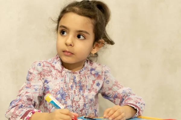 Klein meisje van Perzische verschijning tekent zittend aan het bureau. Mooi meisje van 4 jaar tekent met een waterpotlood. Perzisch meisje lacht — Stockfoto