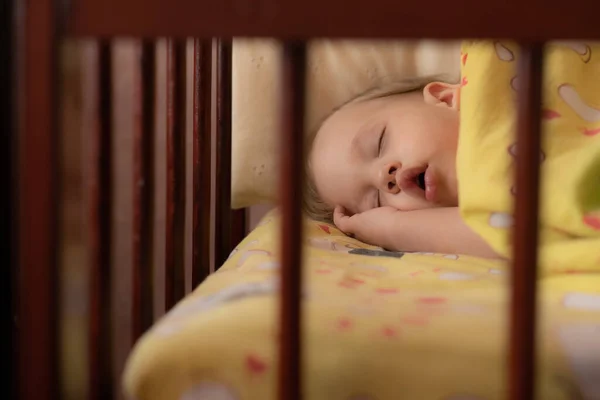 Een jongetje van twee slaapt alleen in een kinderbedje. De baby slaapt in de wieg met zijn hand onder zijn hoofd — Stockfoto
