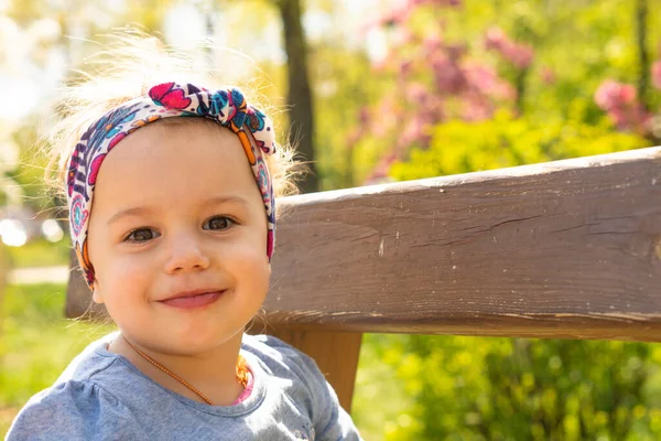 Tijd buiten doorbrengen. Oprechte emoties van een klein meisje. Mooi klein meisje met hoofdband in het voorjaar in het park. close-up uitzicht op kleine meisje die zit op een park bank. — Stockfoto