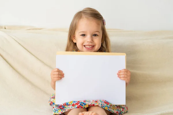 Kleines lächelndes Mädchen mit weißem Blatt Papier Platz für Text, Kopierraum drinnen. Muttertag, Happy Birthday, Zeichnung, Home Art Konzept. — Stockfoto
