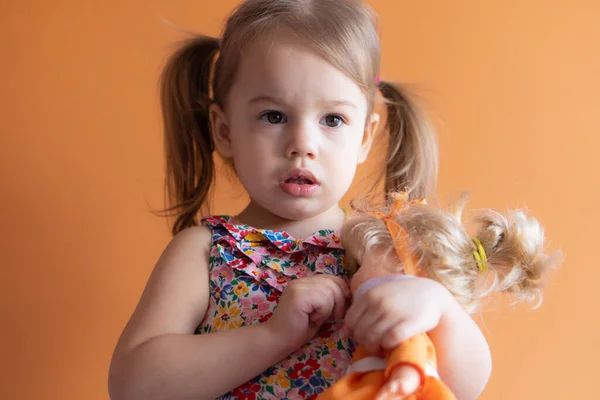 Porträtt av en vacker liten flicka på tre år med orange bakgrund. Barn leker med dockan — Stockfoto