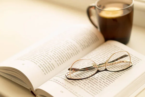 Isolation, loneliness, solitude, poetry, bookbinder, tea party concept. Black mug of warm tea with lemon and open book on the windowsill. Glasses on the opened book