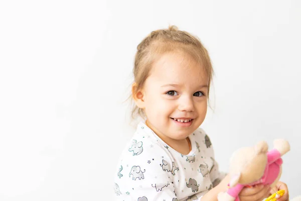 Κοριτσάκι ντυμένο με λευκά ρούχα παίζοντας με παιχνίδια που κάθονται στο κρεβάτι σε εσωτερικούς χώρους. Όμορφο κοριτσάκι με σλαβική εμφάνιση κοιτάζει νέα παιχνίδια. — Φωτογραφία Αρχείου