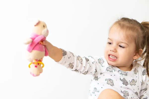 Bambina vestita con abiti bianchi che gioca con i giocattoli seduti sul letto al chiuso. Bella bambina di aspetto slavo sta guardando nuovi giocattoli — Foto Stock