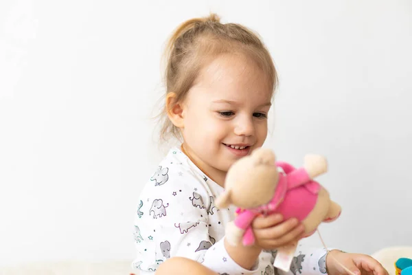 Маленькая девочка в белой одежде играет с игрушками, сидя на кровати в помещении. Красивая маленькая девочка славянской внешности смотрит на новые игрушки — стоковое фото