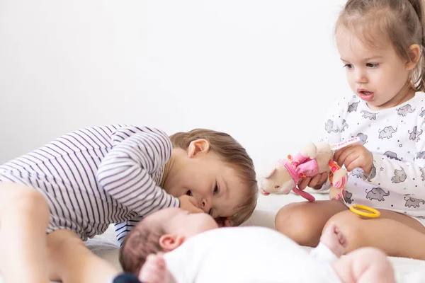 Little smiling kids playing together sitting on the bed. Brother and sister show a newborn a toy. Toddler kids meeting new born sibling. Infant sleeping in white bouncer under a blanket. Kids playing — Stock Photo, Image