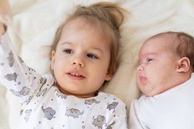 Yatakta birlikte oynayan gülümseyen küçük çocuklar. Erkek ve kız kardeş yeni doğmuş bir bebeğe oyuncak gösterir. Küçük çocuklar yeni doğan kardeşleriyle tanışıyor. Beyaz bir fedaide battaniyenin altında uyuyan bir bebek. Oyun oynayan çocuklar