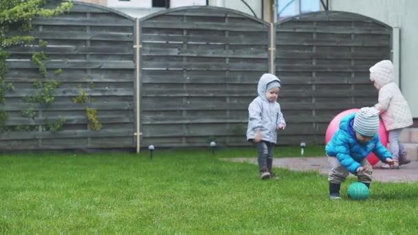 Kindheit, Freizeit, Spiel, Hof, Frühlingskonzept - drei kleine Kinder spielen im Hof bei kaltem Wetter mit Bällen und einem Fitball auf einer grünen Wiese. — Stockvideo