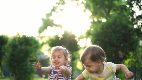 Barndom, familj, livsstil, underhållning och människor koncept - två små barn i samma ålder pojke och flicka riva gräs och spy i parken mot bakgrund av solnedgången. slow motion. — Stockvideo