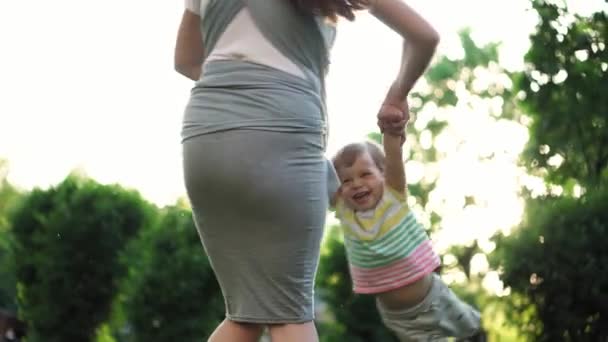 Família, crianças, maternidade, entretenimento e conceito de pessoas - jovem mãe bonito de muitas crianças com seu bebê na funda balançando criança por braços seu filho no fundo do pôr do sol no parque . — Vídeo de Stock
