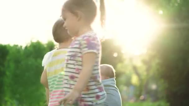 Família, crianças, maternidade, entretenimento e conceito de pessoas - jovem mãe bonito de muitas crianças com seu bebê na funda brinca com duas crianças mais velhas da mesma idade no fundo do pôr do sol no parque . — Vídeo de Stock
