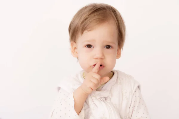 Ένα αγοράκι 2 ετών σε υπνόσακο δείχνει μια χειρονομία να κρατάει αθόρυβα ένα δάχτυλο στα χείλη του στο κρεβάτι. Ένα όμορφο αγόρι με πιτζάμες κάθεται στο κρεβάτι και χαμογελάει. Έννοια των γλυκών ονείρων, οικογένεια — Φωτογραφία Αρχείου