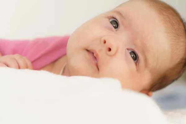 Um recém-nascido deitado de barriga em cima de uma cama branca. O recém-nascido está acordado olhando ao redor na sala. Bebê recém-nascido em um terno rosa sorrindo. Olhar curioso do recém-nascido — Fotografia de Stock