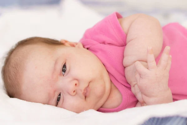 Recién nacida acostada boca abajo en una cama blanca. El recién nacido está despierto mirando a su alrededor en la habitación. Bebé recién nacido en un traje de cuerpo rosa sonriendo. Mirada curiosa del recién nacido —  Fotos de Stock