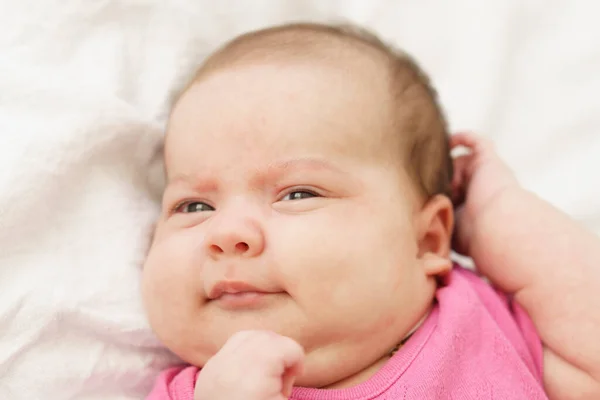 Um recém-nascido deitado de barriga em cima de uma cama branca. O recém-nascido está acordado olhando ao redor na sala. Bebê recém-nascido em um terno rosa sorrindo. Olhar curioso do recém-nascido — Fotografia de Stock