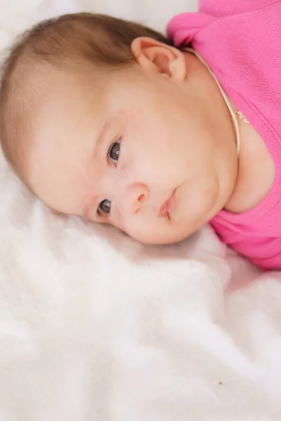 Um recém-nascido deitado de barriga em cima de uma cama branca. O recém-nascido está acordado olhando ao redor na sala. Bebê recém-nascido em um terno rosa sorrindo. Olhar curioso do recém-nascido — Fotografia de Stock
