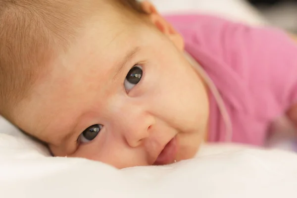 Um recém-nascido deitado de barriga em cima de uma cama branca. O recém-nascido está acordado olhando ao redor na sala. Bebê recém-nascido em um terno rosa sorrindo. Olhar curioso do recém-nascido — Fotografia de Stock