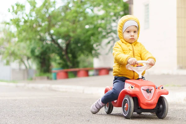 Lieve peuter jongen in een oranje jasje paardrijden plastic speelgoedauto op straat in rustige buurt in het voorjaar bij koud weer — Stockfoto