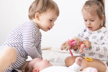 Yatakta birlikte oynayan gülümseyen küçük çocuklar. Erkek ve kız kardeş yeni doğmuş bir bebeğe oyuncak gösterir. Küçük çocuklar yeni doğan kardeşleriyle tanışıyor. Beyaz bir fedaide battaniyenin altında uyuyan bir bebek. Oyun oynayan çocuklar