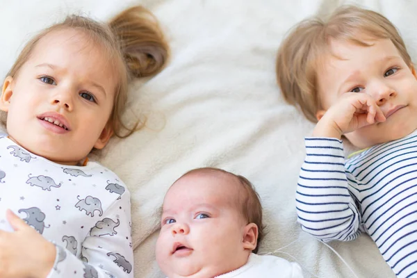 Маленькие улыбающиеся дети, играющие вместе, сидя на кровати. Брат и сестра показывают новорожденному игрушку. Маленькие дети знакомятся с новорожденными братьями и сестрами. Младенец спит в белом вышибале под одеялом. Дети играют — стоковое фото