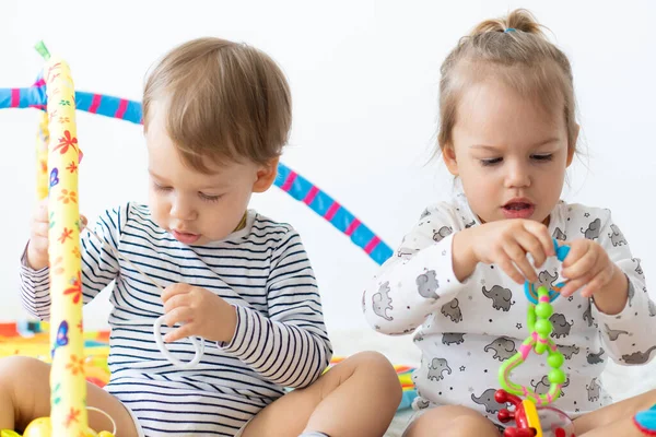 Dos niños pequeños juegan con juguetes juntos sentados en la cama. Hermano y hermana juegan con juguetes de colores. Los niños con una pequeña diferencia de edad juegan juntos en el interior. Juguetes musicales que desarrollan motor —  Fotos de Stock