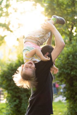 Babalık, ebeveynlik, çocukluk, şefkat, yaz ve eğlence konsepti - sakallı ve siyah tişörtlü genç baba kollarında küçük oğlunu parktaki günbatımının arka ışığında tutuyor..