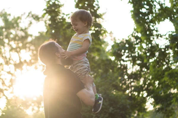 Paternidade, paternidade, infância, carinho, verão e lazer conceito - jovem pai com barba e cabelos longos em t-shirt preta levanta o filho em seus braços na luz de fundo do pôr do sol no parque . — Fotografia de Stock