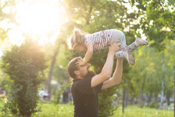 Vaderschap, ouderschap, kindertijd, zorg, zomer en vrije tijd concept - jonge vader met baard en lang haar in zwart t-shirt houdt zijn dochtertje in zijn armen in het licht van de zonsondergang in het park. — Stockfoto