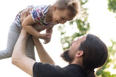 Babalık, ebeveynlik, çocukluk, şefkat, yaz ve eğlence konsepti - sakallı ve siyah tişörtlü genç baba, parkta gün batımının ışığında küçük kızını kollarında tutuyor..