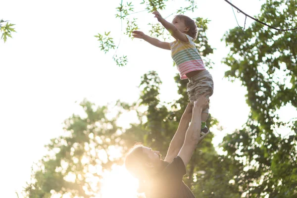 Paternidade, paternidade, infância, carinho, verão e lazer conceito - jovem pai com barba e cabelos longos em t-shirt preta levanta o filho em seus braços na luz de fundo do pôr do sol no parque . — Fotografia de Stock