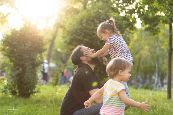 Vaderschap, ouderschap, kindertijd, zorg, zomer en vrije tijd concept - jonge vader met baard en lang haar in zwart t-shirt houdt zijn dochtertje in zijn armen in het licht van de zonsondergang in het park. — Stockfoto