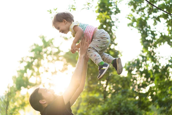 Paternidade, paternidade, infância, carinho, conceito de verão e lazer - o jovem pai com barba e cabelos longos em t-shirt preta joga o seu pequeno filho na luz de fundo do pôr do sol no parque . — Fotografia de Stock