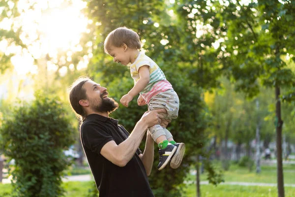 Paternidade, paternidade, infância, carinho, conceito de verão e lazer - o jovem pai com barba e cabelos longos em camiseta preta segura em seus braços o filhinho na luz de fundo do pôr do sol no parque. — Fotografia de Stock