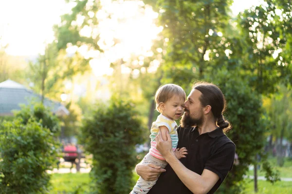 La paternità, la genitorialità, l'infanzia, la cura, l'estate e il tempo libero concetto - giovane papà con la barba e i capelli lunghi in t-shirt nera tiene tra le braccia piccolo figlio nella retroilluminazione del tramonto nel parco. — Foto Stock