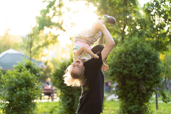 Vaderschap, ouderschap, kindertijd, zorg, zomer en vrije tijd concept - jonge vader met baard en lang haar in zwart t-shirt houdt in zijn armen kleine zoon in de achtergrond van de zonsondergang in het park. — Stockfoto