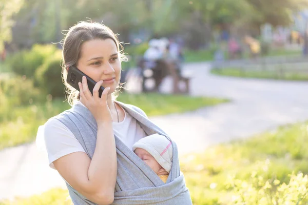 Motherhood, care, infants, summer, communication, parenting concept - Young beautiful mom with a newborn baby in a sling walks and talks on the phone in the backlight of a sunset in the park.