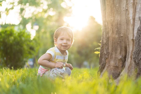 Jeugd, natuur, zomer, parken en buitenleven concept - portret van schattige blonde-harige kleine jongen in gestreepte multi-gekleurde T-shirt glimlachen in de achtergrond van zonsondergang in het park. Een knappe jongen van twee. — Stockfoto