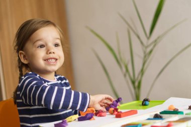 Bebek odasındaki masada plastikle oynayan küçük bir kız. Plastik pervazlar. Bir çocukta iyi motor becerilerin ve yaratıcı hayal gücünün gelişimi. Uzayı kopyala Anaokulu oyunları