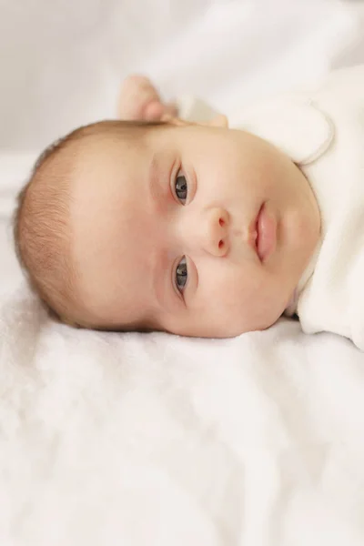 Ένα μικρό νεογέννητο μωρό με λευκό κοστούμι ξαπλωμένο στο κρεβάτι. Κάτοψη ενός νεογέννητου μωρού σε μια λευκή ζεστή κουβέρτα. Το νεογέννητο είναι ξύπνιο και κοιτάζει μέσα. — Φωτογραφία Αρχείου