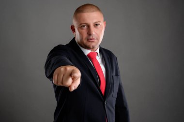 Zarif siyah takım elbiseli, kırmızı kravatlı havalı iş adamı parmağını gri arka planda izole bir şekilde gösteriyor.