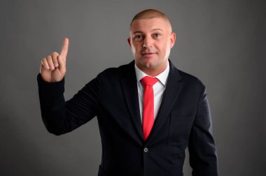 Zarif siyah takım elbiseli ve kırmızı kravatlı havalı iş adamı parmağını gri arka planda izole bir şekilde gösteriyor.