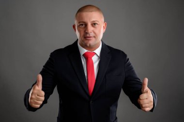 Zarif siyah takım elbiseli ve kırmızı kravatlı havalı iş adamının portresi gri arka planda izole edilmiş çift kamçıları gösteriyor.